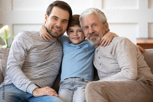Portrait of happy three generations of men sit relax hug on couch in living room. Smiling Caucasian man with small son and elderly father rest on sofa at home show love and care in family relations.