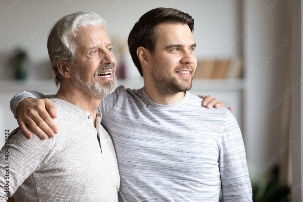 Smiling Caucasian millennial man and senior father hug look in distance dream think of happy future together. Happy grownup son and mature dad embrace visualize and imagine at home. Smiling Caucasian millennial man and senior father hug look in distance dream think of happy future together. Happy grownup son and mature dad embrace visualize and imagine at home.