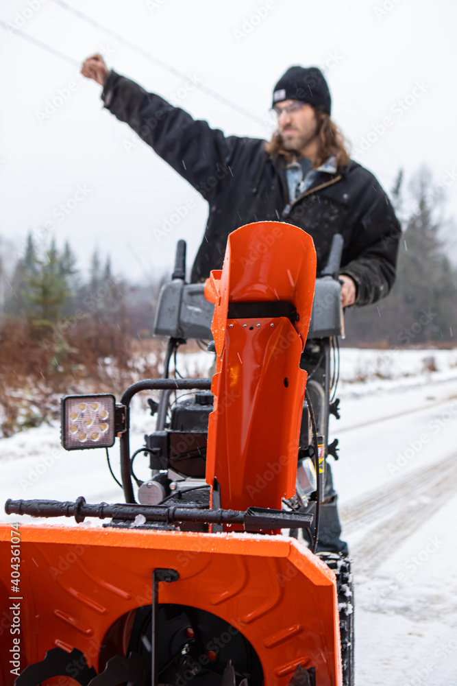 Frontal view of an orange snowplough in a country road in a snowy winter day. Young man in a superhero or superman pose in the blurred background.