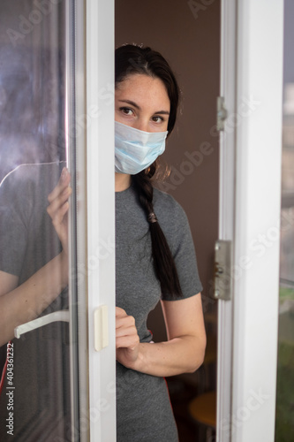 A young brunette girl in a mask with beautiful eyes looks out of the window and looks with hope. Close up. Lockdown, quarantine, epidemic, coronavirus