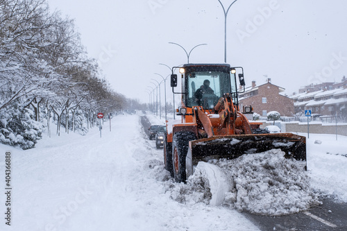A snowplow removes the snow from a street in a residential area, two cars drive behind it. Effects of the Filomena snowstorm