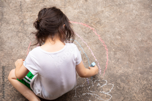 Top view shot of preschool child girl whos is drawing colorful rainbow on pavement or concrete floor in the park by the color chalk in summer time shows the expression of her imagination by activity