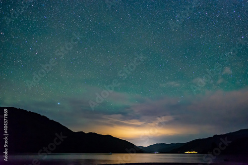 starry sky above the shape of mountains with the light of a city in a fjord in the Marlborough Sounds  New Zealand