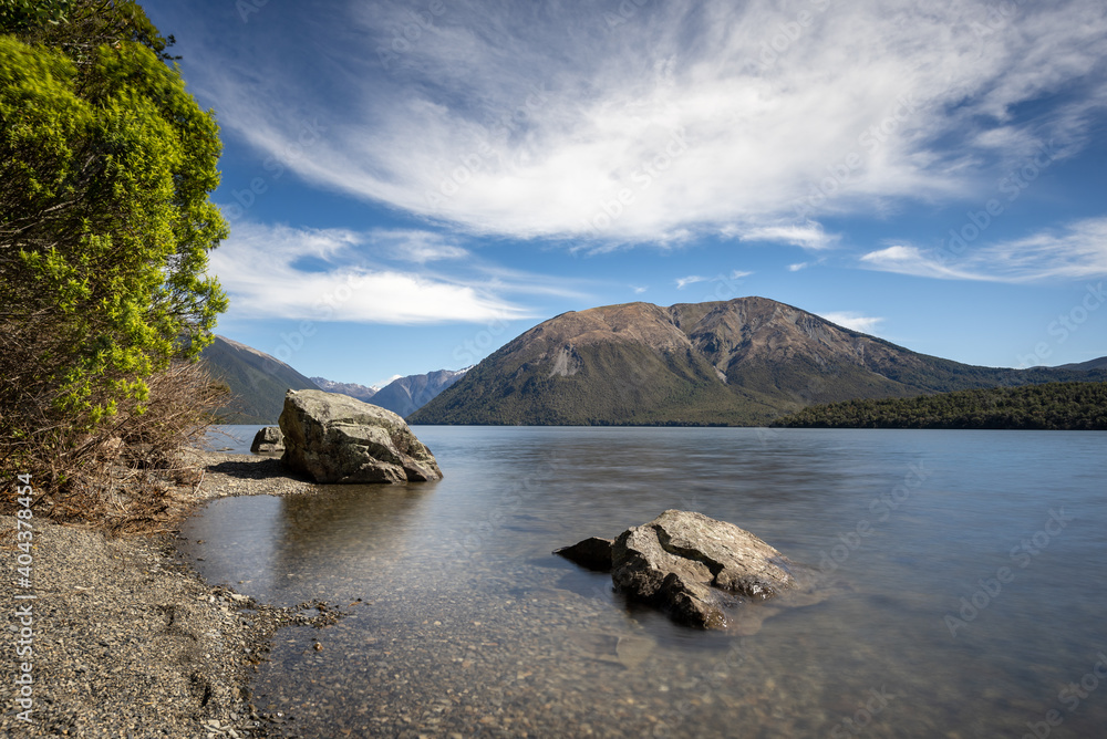 The view across Lake Rotoiti to Pourangahau/Mount Robert in the Nelson Lakes National Park, New Zealand. The shore, trees and two large rocks in the foreground.