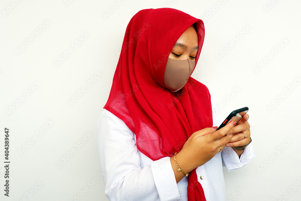 Hijab woman wearing masker looking for something on her smartphone isolated on white background. Hijab woman wearing masker looking for something on her smartphone isolated on white background.