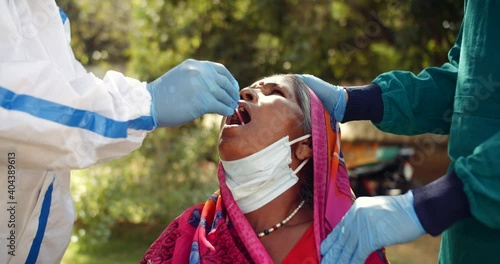 Close-up slow-motion of medical staff with medical gloves takes samples from inside throat of an old woman in red colorful saree for virus examination collection with protective workwear gear in India