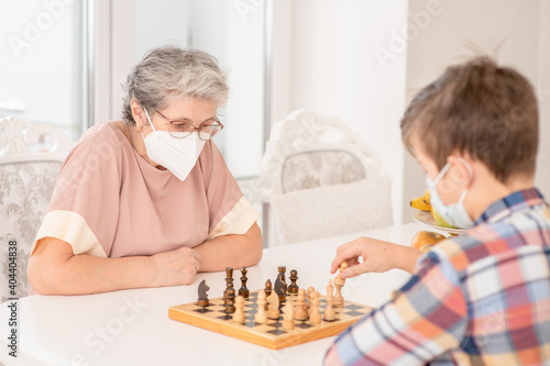 Senior grandmother and her grandson wearing protective face masks play chess at home during quarantine Coronavirus (Covid-19) epidemic