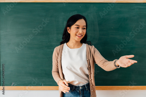 Cheerful young Asian woman gesticulating and looking away while standing near chalkboard during lesson in school © twinsterphoto