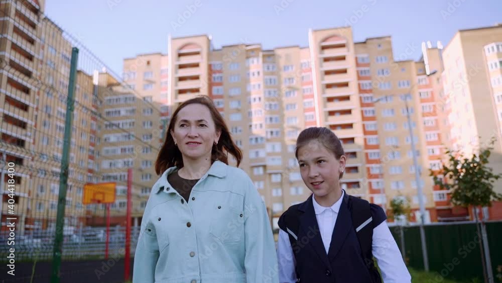 A girl in uniform with a bag on her back, walking next to her mother hand in hand. Mom and daughter are walking down the street to school. A girl in uniform with a bag on her back, walking next to her mother hand in hand. Mom and daughter are walking down the street to school.