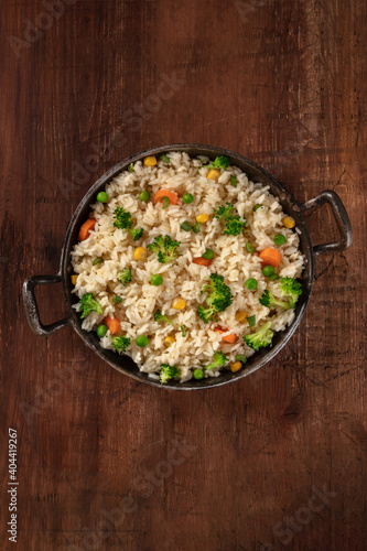 Vegan rice with broccoli, carrots and green peas, shot from above in a frying pan on a dark rustic wooden background, with a place for text