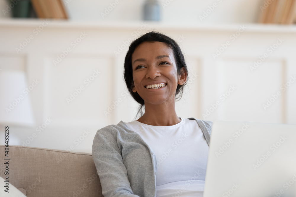 Head shot happy dreamy young african ethnicity woman sitting on couch with laptop, looking in distance. Smiling millennial mixed race freelance worker distracted from doing online job at home. Head shot happy dreamy young african ethnicity woman sitting on couch with laptop, looking in distance. Smiling millennial mixed race freelance worker distracted from doing online job at home.