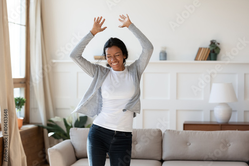 Happy laughing young african american biracial woman dancing to energetic disco music in modern living room, enjoying carefree domestic hobby activity, celebrating freedom on weekend alone indoors.