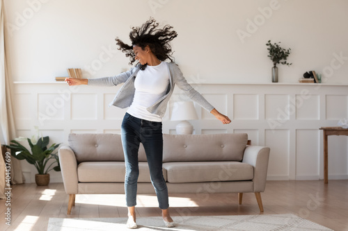 Full length young african ethnicity multiracial woman in casual wear dancing to favorite energetic audio music, having fun alone in living room, enjoying active domestic hobby pastime, feeling freedom photo