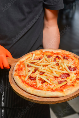 Whole pizza on wooden board. Waiter serving pizza with french fries on top in restaurant or diner, eating out concept