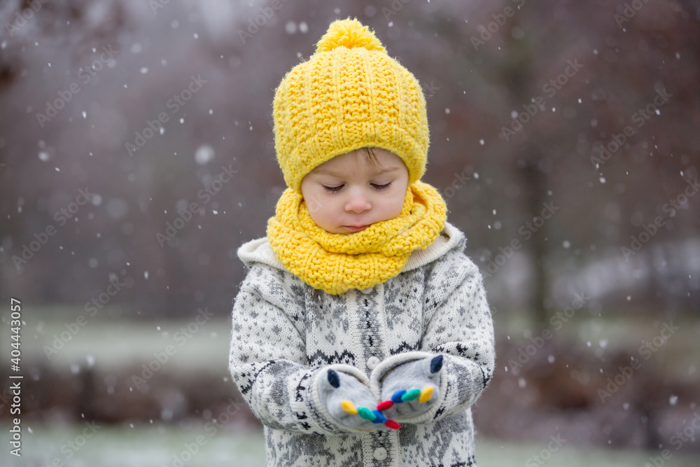 Beautiful blond toddler child, boy, with handmade knitted sweater playing in the park with first snow Beautiful blond toddler child, boy, with handmade knitted sweater playing in the park with first snow
