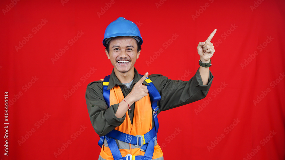 Asian worker man wear unifrom, helmet, safety harness pointing to right blank space isolated red background