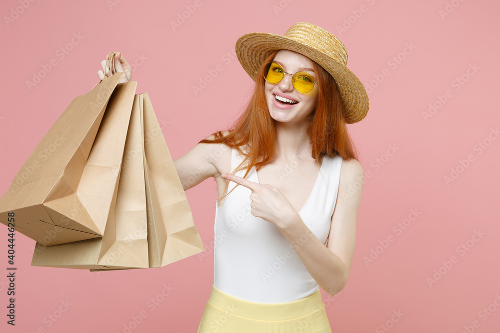 Young smiling redhead happy ginger woman 20s wear straw hat glasses summer clothes hold point finger on package bags with purchases after shopping isolated on pastel pink background studio portrait Young smiling redhead happy ginger woman 20s wear straw hat glasses summer clothes hold point finger on package bags with purchases after shopping isolated on pastel pink background studio portrait