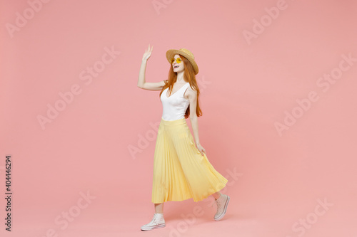 Full length body young smiling redhead ginger woman in straw hat glasses summer clothes maxi skirt walk going waving hand greet someone look aside isolated on pastel pink background studio portrait