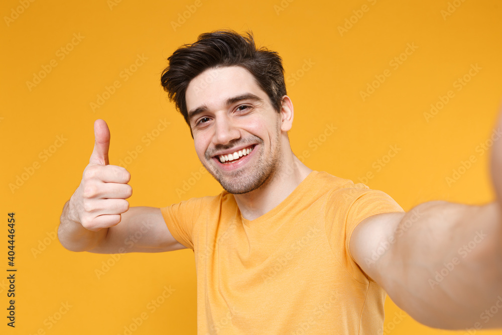 Close up young fun smiling unshaved friendly caucasian handsome man wearing basic blank t-shirt doing selfie shot on mobile phone show thumbs up isolated on yellow color background studio portrait Close up young fun smiling unshaved friendly caucasian handsome man wearing basic blank t-shirt doing selfie shot on mobile phone show thumbs up isolated on yellow color background studio portrait
