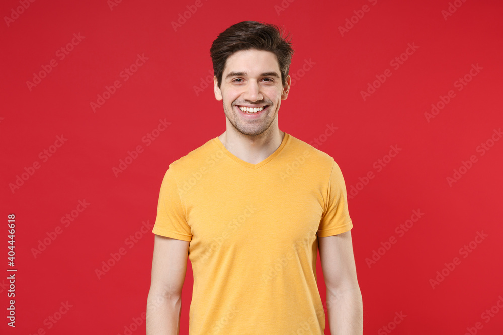 Fototapeta premium Smiling young unshaved caucasian friendly cheerful happy man 20s years old wearing casual basic blank print design yellow t-shirt looking camera isolated on red color background studio portrait.