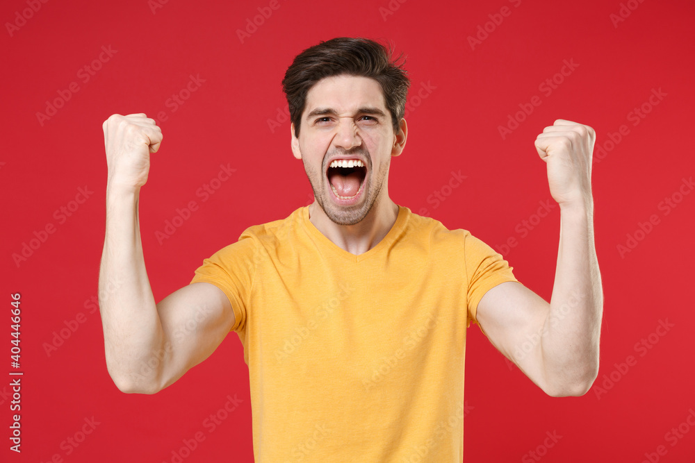 Φωτογραφία Young smiling excited successful unshaved caucasian handsome man 20s wearing casual design yellow t-shirt doing winner gesture with clenching fists isolated on red color background studio portrait