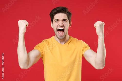 Young smiling excited successful unshaved caucasian handsome man 20s wearing casual design yellow t-shirt doing winner gesture with clenching fists isolated on red color background studio portrait.