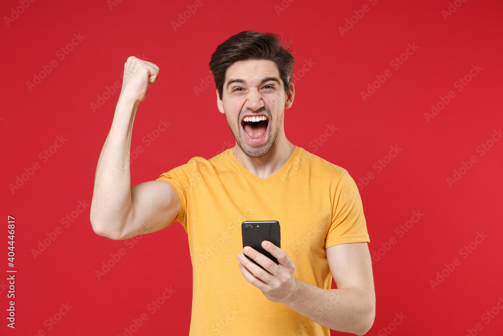 Young surprised unshaved excited man 20s in casual basic yellow t-shirt holding modern mobile cell phone doing winner gesture saying yes clenching fists isolated on red background studio portrait. Young surprised unshaved excited man 20s in casual basic yellow t-shirt holding modern mobile cell phone doing winner gesture saying yes clenching fists isolated on red background studio portrait.