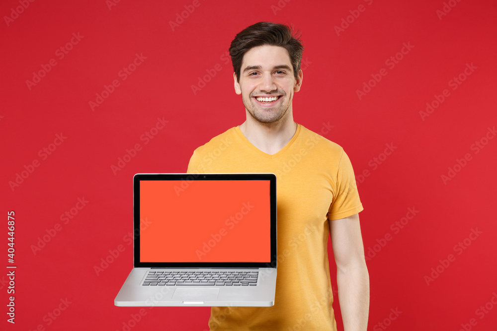 Young smiling unshaved caucasian happy man 20s in yellow t-shirt using holding laptop pc computer with blank screen workspace area copy space mock up isolated on red color background studio portrait. Young smiling unshaved caucasian happy man 20s in yellow t-shirt using holding laptop pc computer with blank screen workspace area copy space mock up isolated on red color background studio portrait.