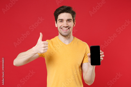 Young satisfied happy man 20s in casual yellow t-shirt hold mobile cell phone with blank screen workspace copy space mock up showing thumb up gesture isolated on red color background studio portrait.