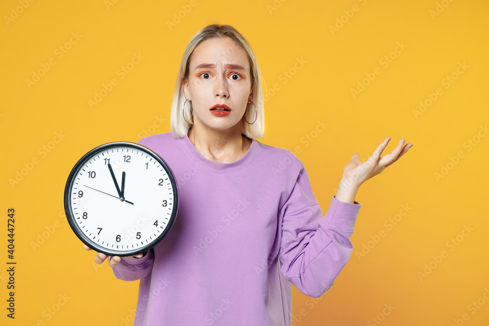 Young blonde caucasian shocked confused woman 20s with bob haircut makeup wearing casual basic purple shirt with spreading hand holding in hands clock isolated on yellow background studio portrait. Young blonde caucasian shocked confused woman 20s with bob haircut makeup wearing casual basic purple shirt with spreading hand holding in hands clock isolated on yellow background studio portrait.