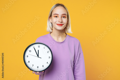 Young blonde caucasian beautiful woman 20s with bob haircut bright makeup wearing casual basic purple shirt holding in hands clock showing time isolated on yellow color background studio portrait.