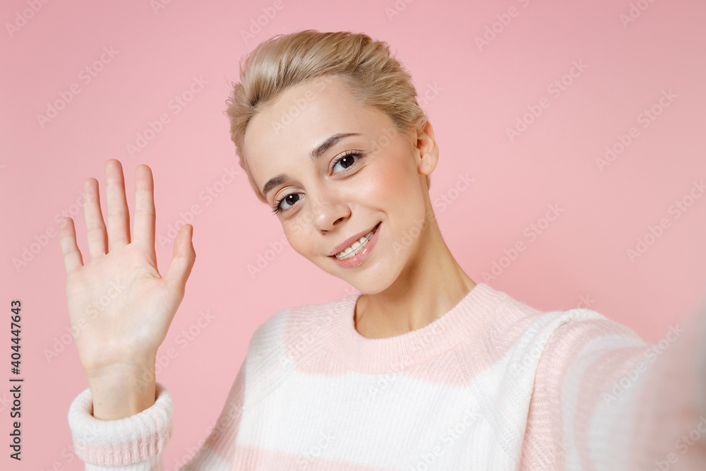 Close up of young smiling woman short haircut nude makeup dental braces in sweater do selfie shot on mobile phone greet with hand as notices someone isolated on pastel pink background studio portrait Close up of young smiling woman short haircut nude makeup dental braces in sweater do selfie shot on mobile phone greet with hand as notices someone isolated on pastel pink background studio portrait
