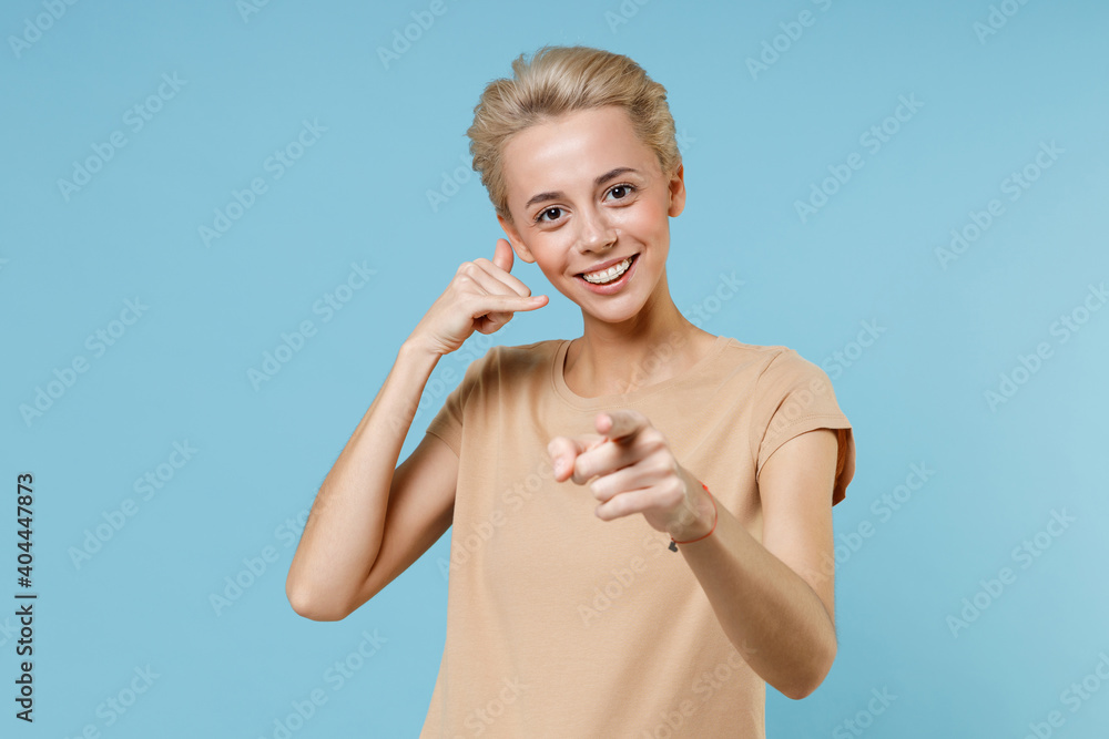 Young blonde smiling woman dental braces short haircut in casual beige t-shirt doing phone gesture like says call me back point index finger camera on you isolated on blue background studio portrait Young blonde smiling woman dental braces short haircut in casual beige t-shirt doing phone gesture like says call me back point index finger camera on you isolated on blue background studio portrait
