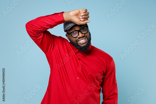 Exhausted tired young bearded african american man 20s wearing casual red shirt eyeglasses cap standing put hand on head looking camera isolated on pastel blue color wall background studio portrait. photo