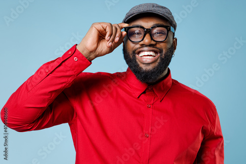 Leinwand Poster Close up of laughing cheerful young bearded african american man 20s wearing casual red shirt cap eyeglasses standing and looking camera isolated on pastel blue color wall background studio portrait