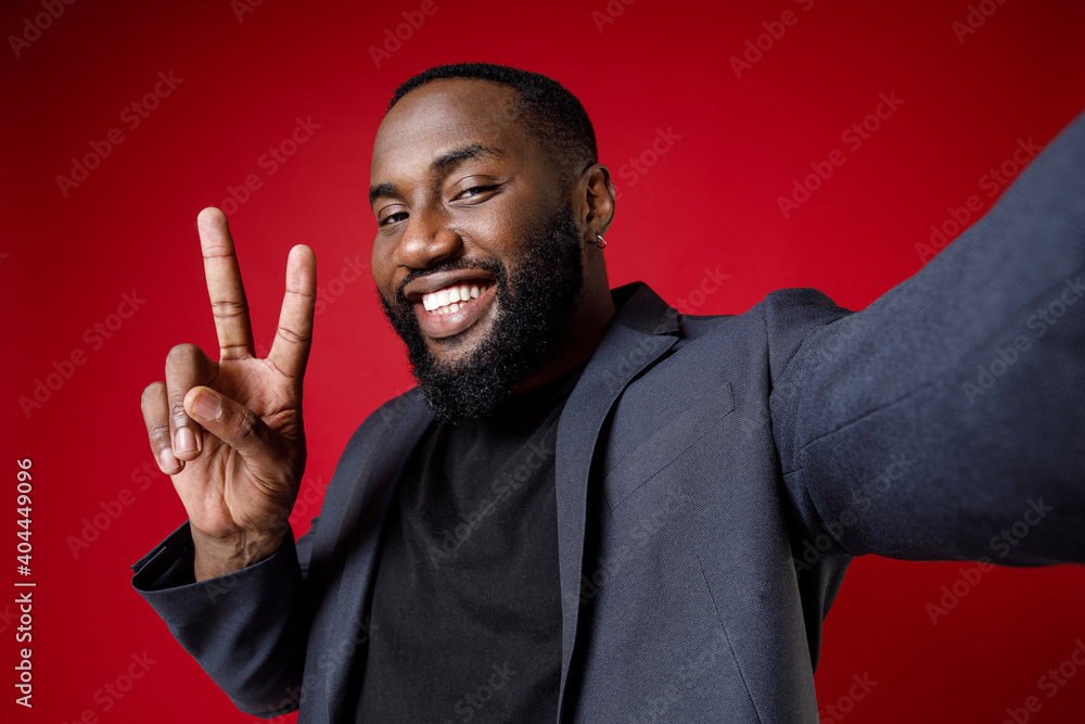 Close up of smiling young african american business man 20s in classic jacket suit standing doing selfie shot on mobile phone showing victory sign isolated on red color background studio portrait. Close up of smiling young african american business man 20s in classic jacket suit standing doing selfie shot on mobile phone showing victory sign isolated on red color background studio portrait.