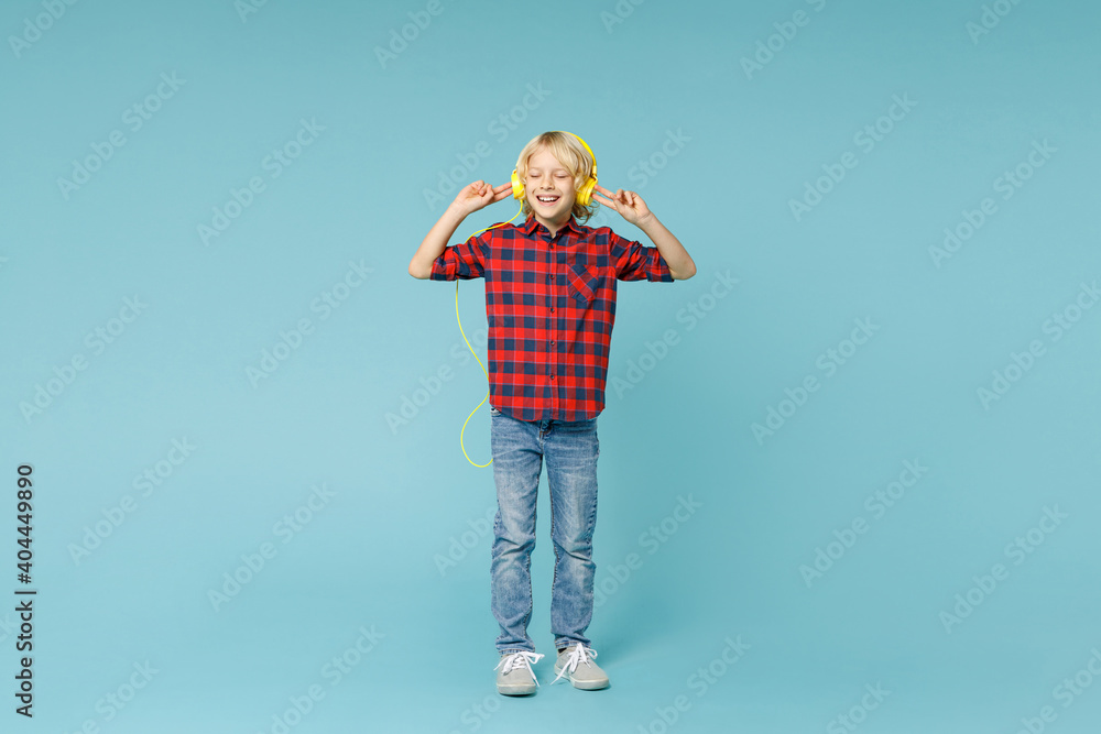 Full length of smiling little curly kid boy 10s in basic red checkered shirt listening music with headphones isolated on blue color background children studio portrait. Childhood lifestyle concept. Full length of smiling little curly kid boy 10s in basic red checkered shirt listening music with headphones isolated on blue color background children studio portrait. Childhood lifestyle concept.