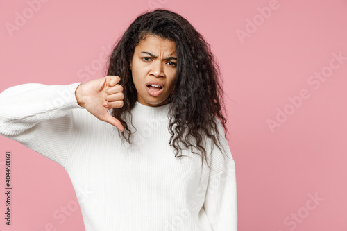 Young disgusted ispleased african american woman 20s curly hair in white knitted sweater look camera showing thumb down dislike isolated on pastel pink background studio portrait. Lifestyle concept. photo