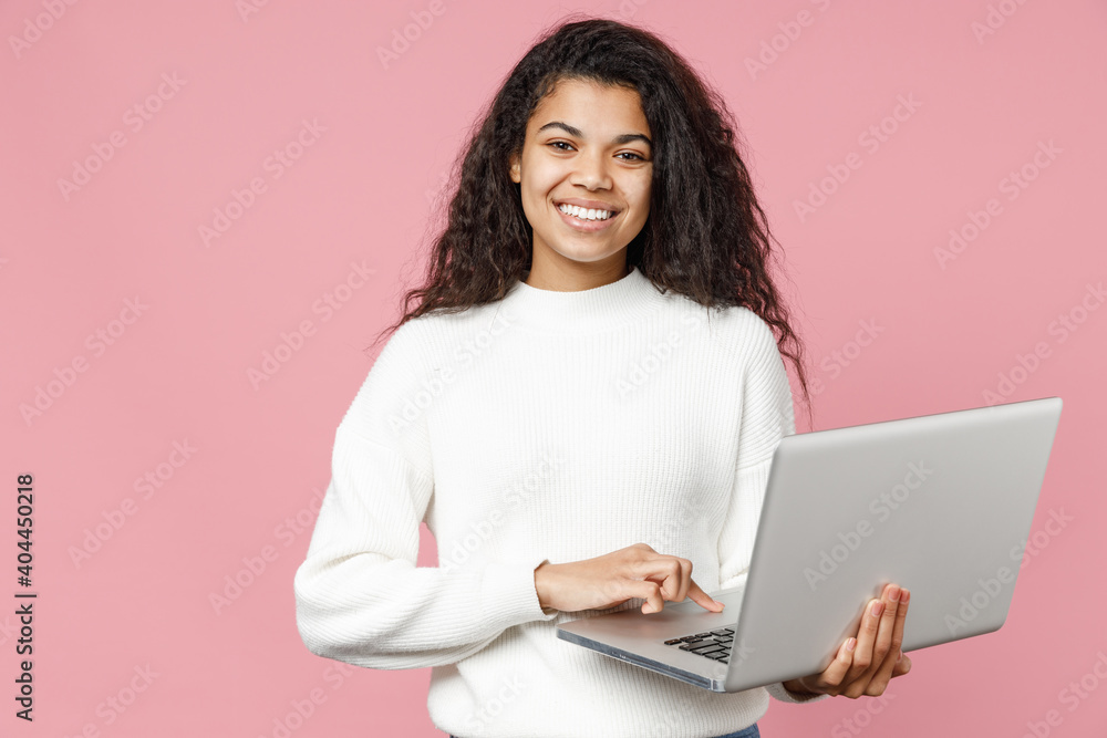 Young smiling african american woman 20s curly hair wear white casual knitted sweater hold laptop pc computer typing chat on keyboard look camera isolated on pastel pink background studio portrait Young smiling african american woman 20s curly hair wear white casual knitted sweater hold laptop pc computer typing chat on keyboard look camera isolated on pastel pink background studio portrait