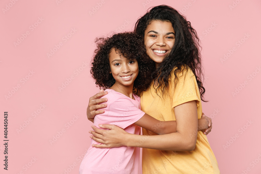 Cheerful smiling african american young woman and little kid girl sisters wearing casual t-shirts hugging looking camera isolated on pastel pink color background studio portrait. Family day concept. Cheerful smiling african american young woman and little kid girl sisters wearing casual t-shirts hugging looking camera isolated on pastel pink color background studio portrait. Family day concept.