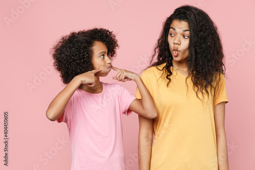 Funny african american young woman little kid girl sisters in t-shirts point fingers on blowing puffing cheeks with funny face mouth inflated with air isolated on pink background. Family day concept.