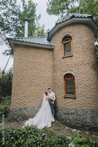 A smiling  curly-haired bride in lacy white and a stylish groom in a blue striped suit are hugging against the backdrop of a brick fortress  castle  walls. Wedding portrait of the newlyweds.