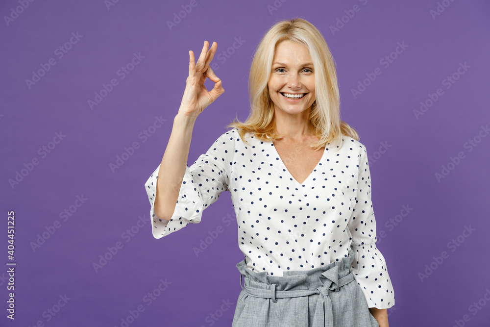 Smiling funny elderly gray-haired blonde woman lady 40s 50s years old in white dotted blouse standing showing OK gesture looking camera isolated on bright violet color background studio portrait. Smiling funny elderly gray-haired blonde woman lady 40s 50s years old in white dotted blouse standing showing OK gesture looking camera isolated on bright violet color background studio portrait.