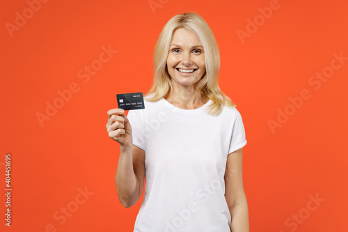 Smiling elderly gray-haired blonde woman lady 40s 50s years old in white basic t-shirt standing hold in hand credit bank card looking camera isolated on bright orange color background studio portrait.