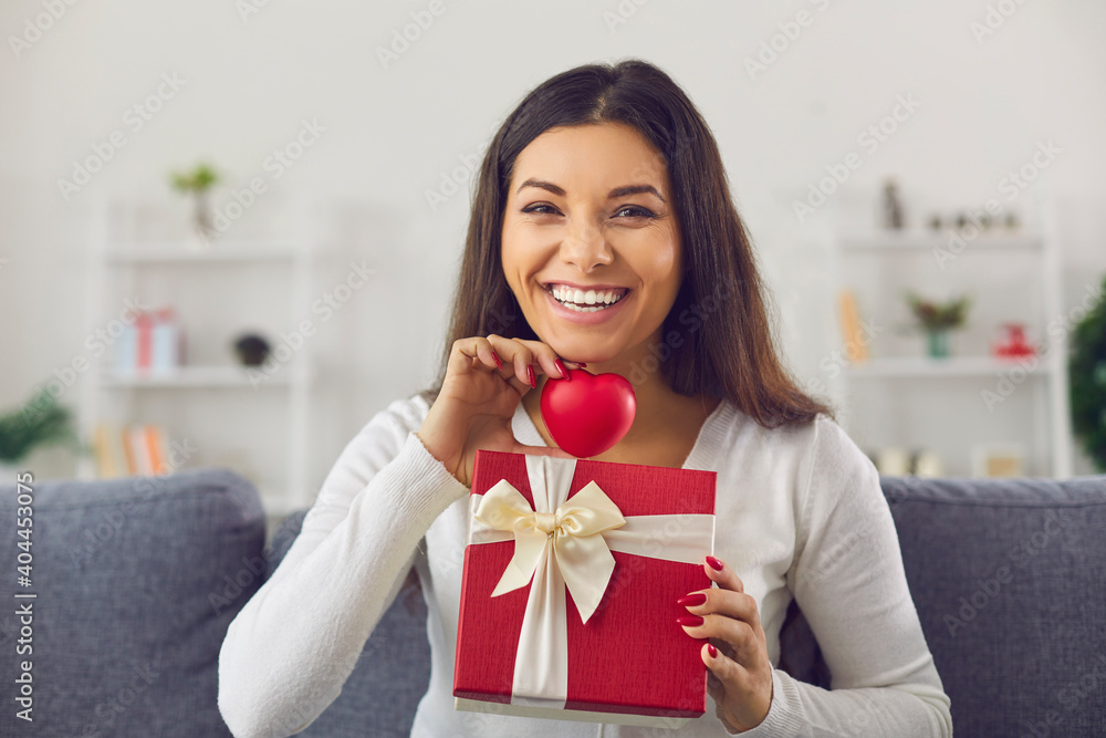 Skype conference headshot happy smiling young woman in love sitting on sofa at home, holding red heart and gift box, saying thank you for Valentine present. Blogger doing giveaway and recording video Skype conference headshot happy smiling young woman in love sitting on sofa at home, holding red heart and gift box, saying thank you for Valentine present. Blogger doing giveaway and recording video