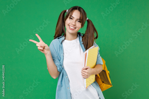 Funny smiling young woman student in basic blue shirt backpack hold notebooks showing victory sign isolated on green background studio portrait. Education in high school university college concept.