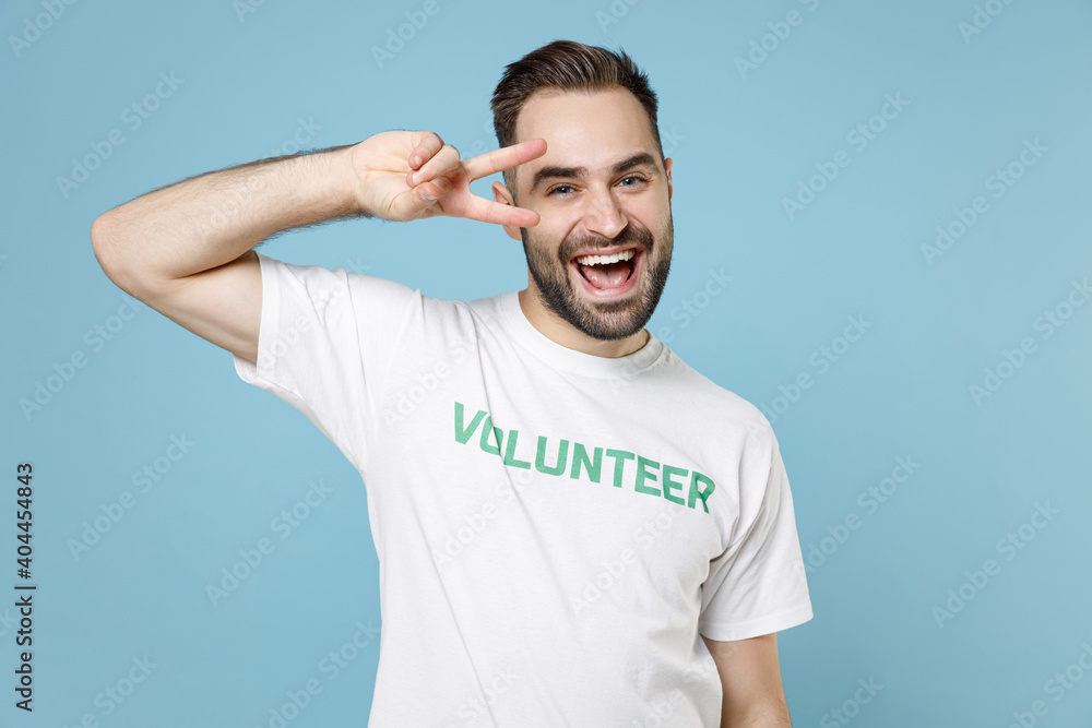 Cheerful young bearded man in white volunteer t-shirt showing victory sign looking camera isolated on blue color background studio portrait. Voluntary free work assistance help charity grace concept. Cheerful young bearded man in white volunteer t-shirt showing victory sign looking camera isolated on blue color background studio portrait. Voluntary free work assistance help charity grace concept.