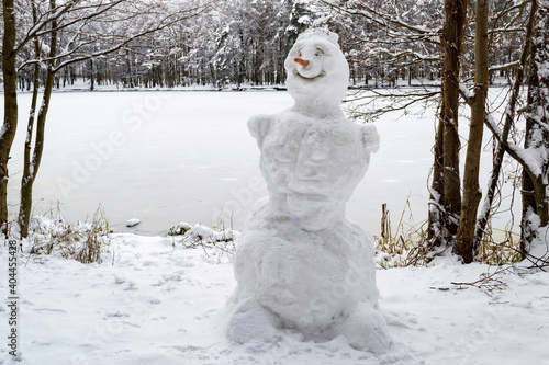 A snowman stands on the shore of the lake. Snowy forest and frozen lake. Concept of winter games and New Year's mood.