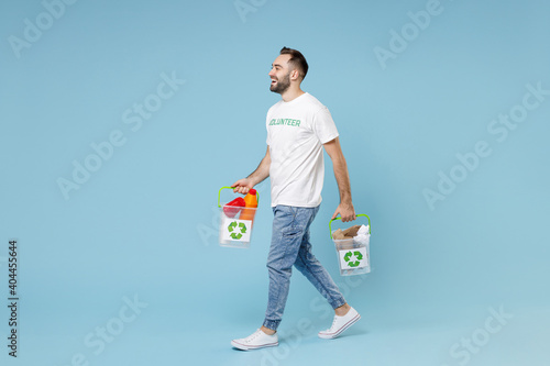 Full length side view of funny young man in volunteer t-shirt hold baskets with plastic paper trash isolated on blue background studio. Voluntary free assistance help trash sorting recycling concept.