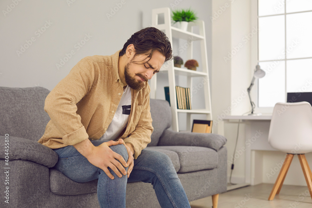 Unhappy man sitting on sofa at home and touching hurt knee with grimace of pain and suffering. Young guy with rheumatism, arthritis or osteoarthritis feeling intense severe pain in knee joint Unhappy man sitting on sofa at home and touching hurt knee with grimace of pain and suffering. Young guy with rheumatism, arthritis or osteoarthritis feeling intense severe pain in knee joint